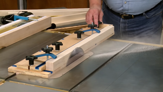 Plywood tapering sled with adjustable blue hold-down clamps securing a wooden leg on a table saw